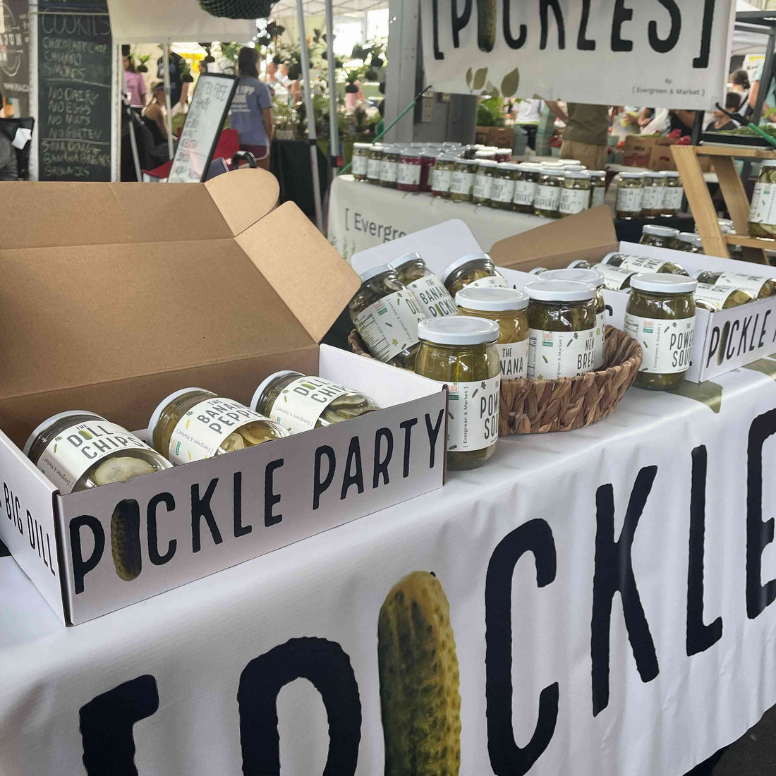 Open Pickle Party box of pickle jars on a market table with a [PICKLES] banner