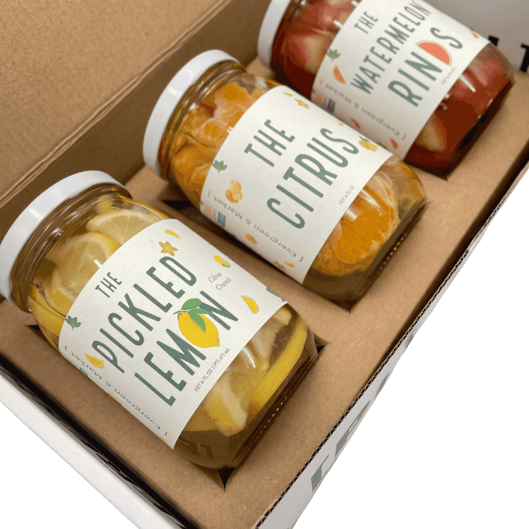 Three jars of pickled fruits and vegetables in a gift box labeled 'The Pickled Lemon' with lemon slices inside, on a brown cardboard tray.