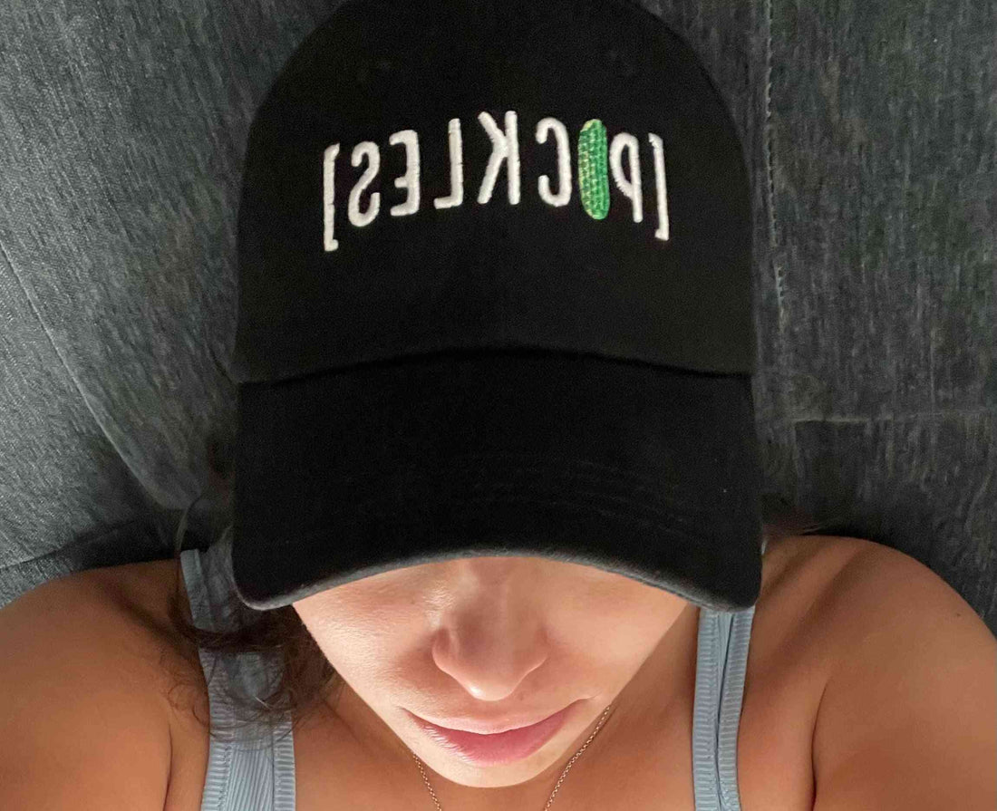 Close-up of black Pickle Cap Hat with embroidered [PICKLES] text and green pickle detail, worn by a person indoors