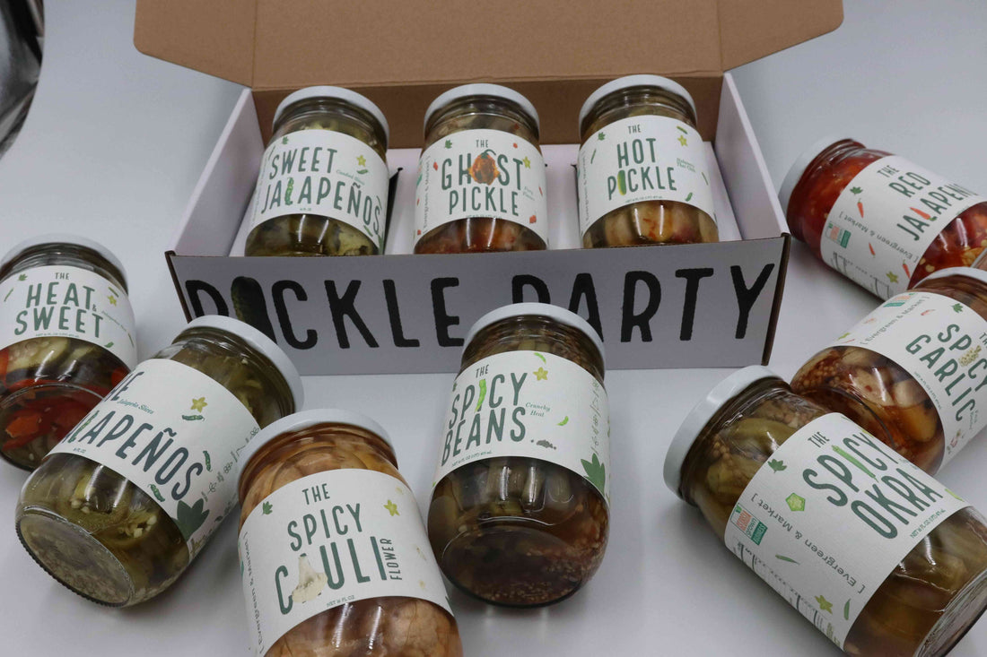 Assorted jars of pickled vegetables with labels like 'Sweet Jalapenos' and 'Ghost Pickle' in a white display box at a market stall.