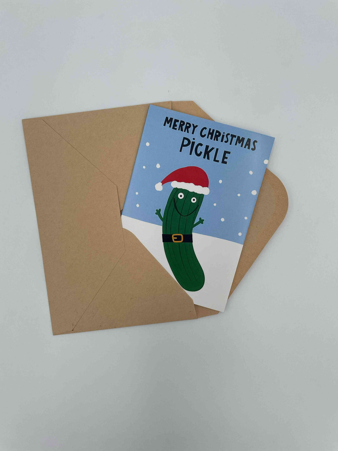 Merry Christmas Pickle card with a pickle in a Santa suit on a blue background, on a kraft envelope